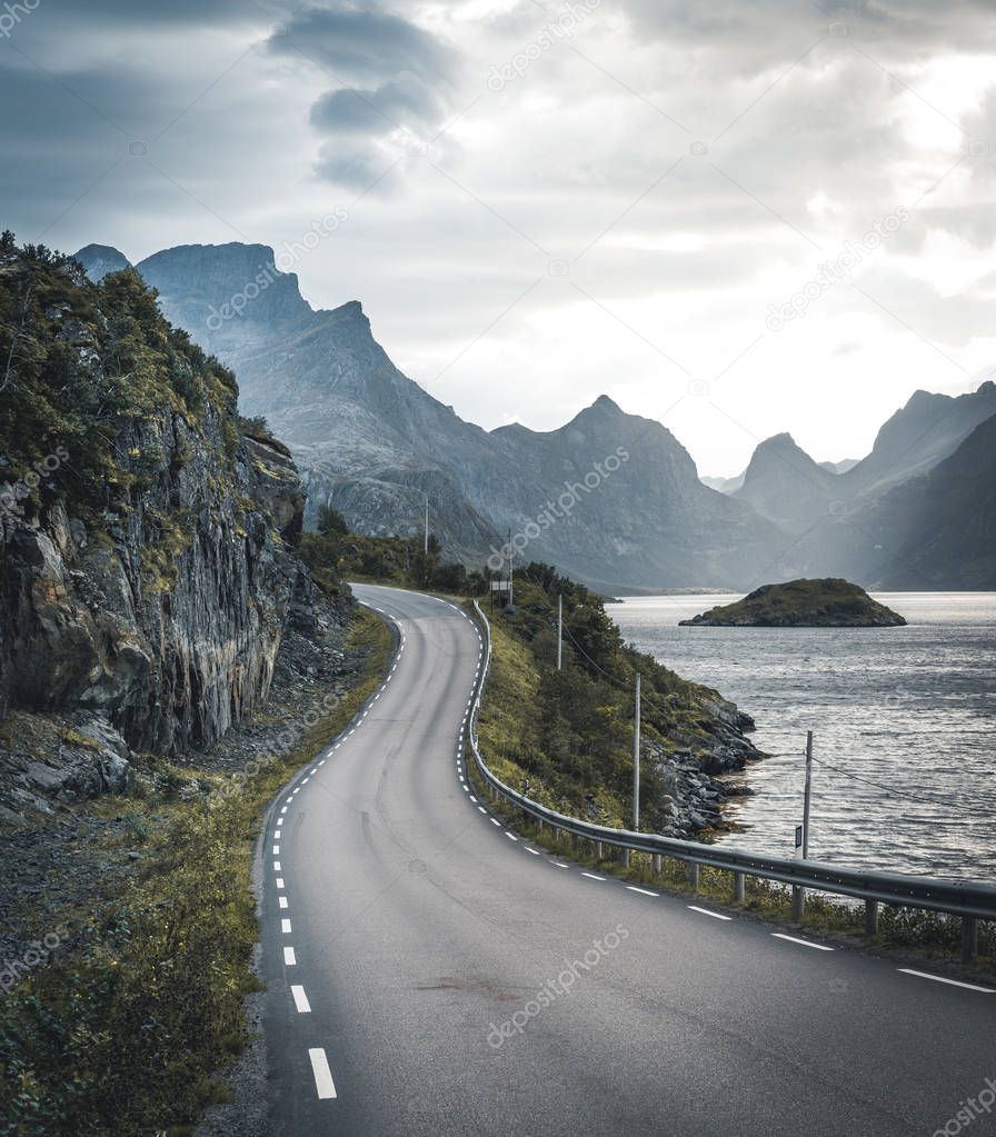 Winding road trip towards Henningsvaer on Lofoten Islands with mountains in the background. Cloudy and moody sky with atlantic ocean.