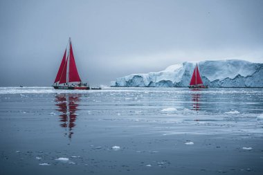 Beautiful red sailboat in the arctic next to a massive iceberg showing the scale. Cruising among floating icebergs in Disko Bay glacier during midnight sun season of polar summer Ilulissat, Disko Bay clipart