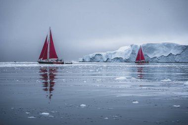 Kuzey kutbunda, ölçeği gösteren devasa bir buzdağının yanında güzel kırmızı yelkenli. Polar yaz Ilulissat, Disko Körfezi gece yarısı güneş sezonu sırasında Disko Körfezi buzul yüzen buzdağları arasında seyir