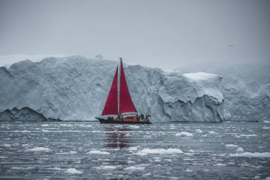 Kuzey kutbunda, ölçeği gösteren devasa bir buzdağının yanında güzel kırmızı yelkenli. Polar yaz Ilulissat, Disko Körfezi gece yarısı güneş sezonu sırasında Disko Körfezi buzul yüzen buzdağları arasında seyir