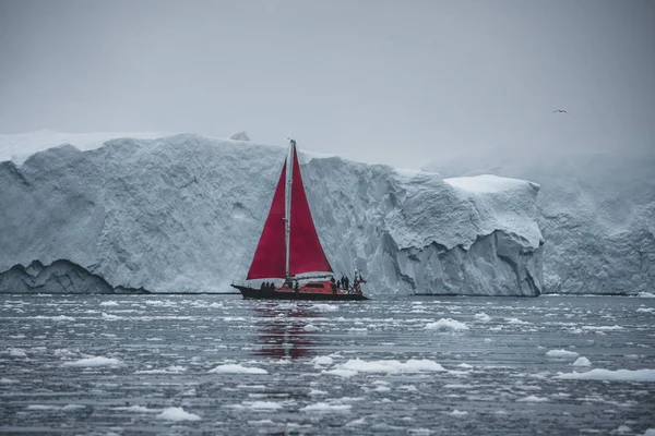 Vacker röd segelbåt i Arktis bredvid ett massivt isberg som visar skalan. Cruising bland flytande isberg i disko Bay Glacier under midnattssol säsongen av Polar Summer Ilulissat, disko Bay — Stockfoto