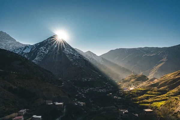 Ein Blick auf den Sonnenuntergang mit Sonnenstern im ländlichen Bergdorf Imlil im Hohen Atlas in Marokko in Afrika. Schneebedeckte Gipfel im Hintergrund mit Leuchtkästen und blauem Himmel. — Stockfoto