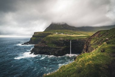 Gasadalur köyü ve Mulafossur 'un ikonik şelalesi Vagar, Faroe Adaları' ndaki hava aracı panoraması. Kuzey Atlantik Okyanusu 'nda zor görünüyor. Yaz boyunca yemyeşil.