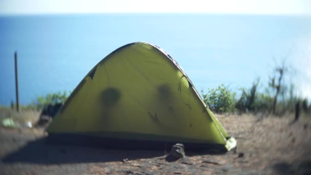 A tourist tent on the seashore on a rocky shore, through which the rays of the sun pass. silhouettes of people talking in a tent. 4k. — Stock Video