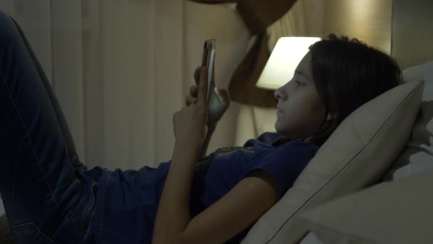 Roztomilá holčička ve svém pokoji v noci, ležící na posteli pomocí smartphonu. její noční lampa svítí. 4k. — Stock video