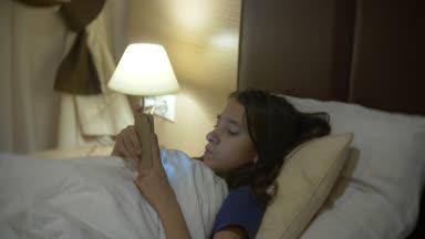 Onun odasına gece, sevimli küçük kız Smartphone kullanarak yatakta yatıyordu. Onun gece lambası yanıyor. 4k.