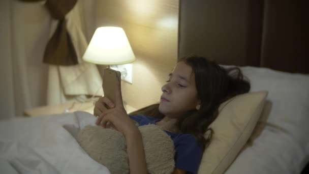 Cute little girl in her room at night, lying on bed using smartphone. her night lamp is on. 4k. — Stock Video