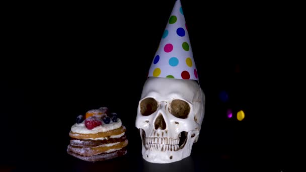 Gâteau et crâne dans une casquette festive sur un fond noir. bonbons, tir au ralenti. 4k — Video