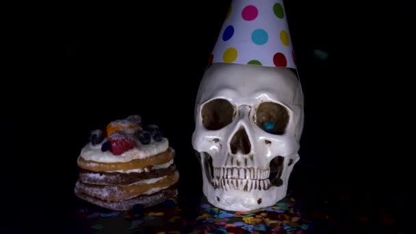 Cake and skull in a festive cap on a black background. 4k, dolly shot, defocusing, blur. — Stock Video