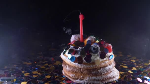 Fiesta, celebración y concepto de fiesta - magdalena de cumpleaños con una vela encendida sobre fondo negro, confeti. Confetti para una fiesta. super cámara lenta — Vídeos de Stock