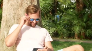 Bir palmiye ağacının altında oturan ve onun smartphone kullanarak yakışıklı bir adam. 4k.