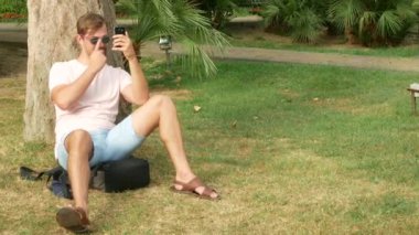 Bir palmiye ağacının altında oturan ve onun smartphone kullanarak yakışıklı bir adam. 4k.