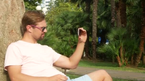 Ένας όμορφος άνδρας κάθεται κάτω από ένα φοίνικα και τη χρήση του smartphone. 4k. — Αρχείο Βίντεο