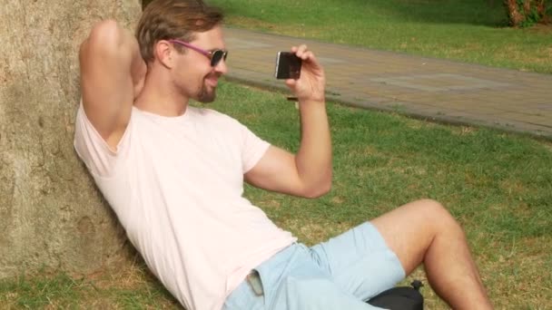 Un hombre guapo sentado debajo de una palmera y usando su teléfono inteligente. 4k . — Vídeos de Stock