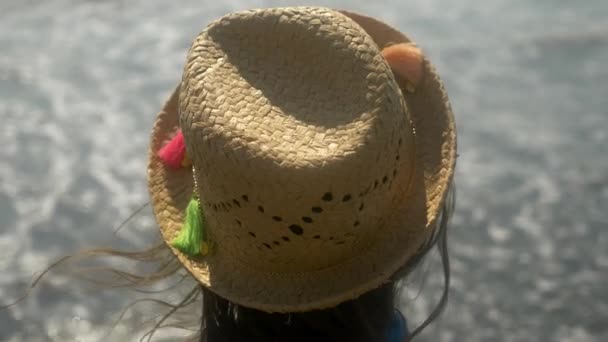 4k, primer plano. la chica en un sombrero de paja mira las olas del mar. Vista trasera. Movimiento lento — Vídeo de stock