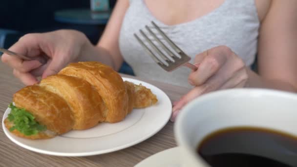 Une femme méconnaissable avec un beau décolleté, des petits déjeuners dans un café avec un croissant sandwich au porc bouilli et boire du café. 4k, ralenti, gros plan . — Video