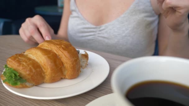 Une femme méconnaissable avec un beau décolleté, des petits déjeuners dans un café avec un croissant sandwich au porc bouilli et boire du café. 4k, ralenti, gros plan . — Video