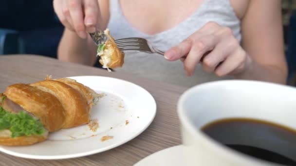 Une femme méconnaissable avec un beau décolleté, des petits déjeuners dans un café avec un croissant sandwich au porc bouilli et boire du café. 4k, ralenti, gros plan . — Video