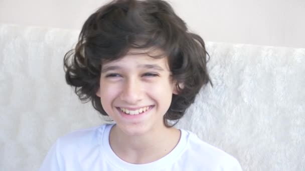 Schattige curly-haired jongen de tiener kijkt naar de camera lachen en maakt een grappige gelaatsuitdrukking. 4 k, slow motion, close-up — Stockvideo