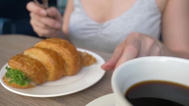 Una mujer irreconocible con un hermoso escote, desayunos en un café con un sándwich de croissant con cerdo hervido y beber café. 4k, cámara lenta, primer plano . — Vídeos de Stock