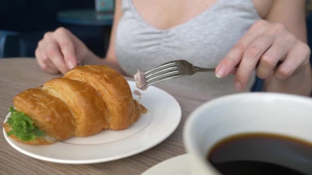 Une femme méconnaissable avec un beau décolleté, des petits déjeuners dans un café avec un croissant sandwich au porc bouilli et boire du café. 4k, ralenti, gros plan . — Video