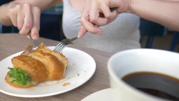 Uma mulher irreconhecível com um belo decote, pequenos-almoços em um café com um sanduíche croissant com carne de porco fervida e beber café. 4k, câmera lenta, close-up . — Vídeo de Stock