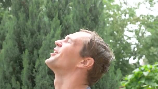 Молодой красивый парень под дождем. капли воды падают ему на лицо. крупный план 4k, замедленная съемка — стоковое видео