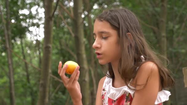 Schattig klein meisje brunette, fruit eten en praten. Achtergrond van de natuur. 4k, slow-motion — Stockvideo
