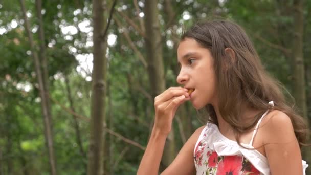 Aranyos kis lány barna, eszik gyümölcsöt és beszél. Természet háttér. 4k, lassú mozgás — Stock videók