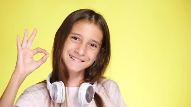 Chica adolescente con auriculares y teléfono móvil escuchando música en el fondo de color. Primer plano, 4k, cámara lenta. muestra gesto, ok, super — Vídeo de stock