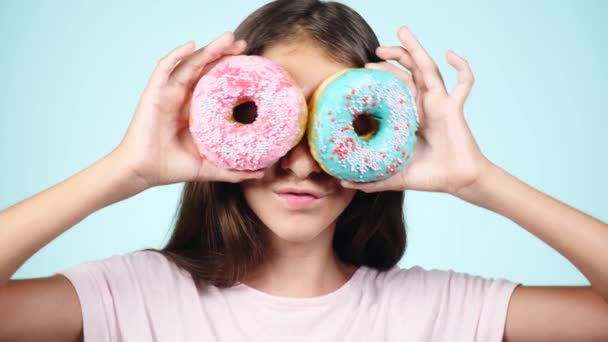 Close-up. Portret van een grappig meisje met lange haren, plezier maken met kleurrijke donuts tegen haar gezicht. Expressies, dieet concept, achtergrondkleur — Stockvideo
