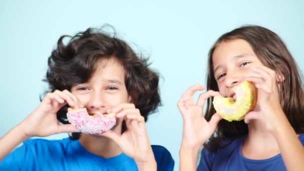 Close-up. retrato de um cara engraçado e uma menina, se divertindo com donuts coloridos em seu rosto. Expressões, conceito de dieta, cor de fundo. 4k — Vídeo de Stock