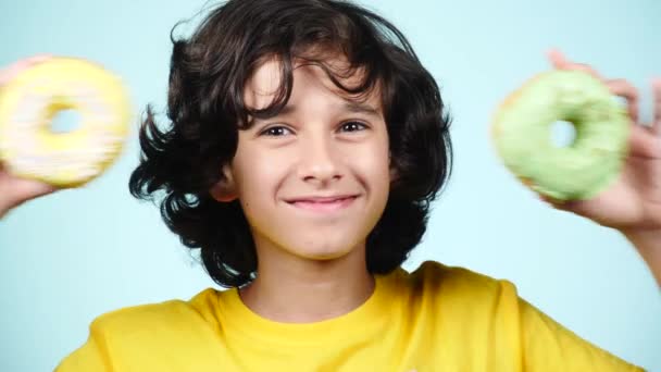 Primo piano. ritratto di un ragazzo divertente con i capelli ricci, divertirsi con ciambelle colorate contro il suo viso. Espressioni, concetto di dieta, colore di sfondo. 4k — Video Stock