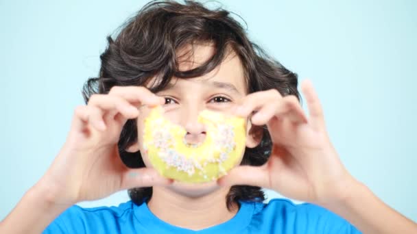 Nahaufnahme. Porträt eines lustigen Typen mit lockigen Haaren, der Spaß mit bunten Donuts vor seinem Gesicht hat. Ausdruck, Ernährungskonzept, Hintergrundfarbe. 4k — Stockvideo