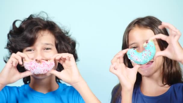 Close-up. retrato de um cara engraçado e uma menina, se divertindo com donuts coloridos em seu rosto. Expressões, conceito de dieta, cor de fundo. 4k — Vídeo de Stock