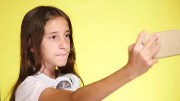 Chica adolescente con auriculares y teléfono móvil escuchando música en el fondo de color. Primer plano, 4k, cámara lenta. hace selfi en el teléfono inteligente — Vídeo de stock