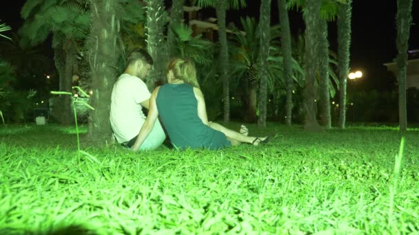 Amoroso casal mulher e homem sentado à noite em um jardim tropical sob uma palmeira. 4K. escuro — Vídeo de Stock