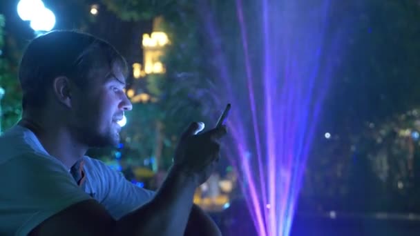Mladý pohledný kluk sedí večer u fontána s osvětlením barevné vody. mluvil po telefonu, rozostření, 4k — Stock video