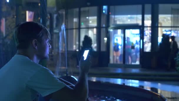 Un chico guapo joven se sienta en la noche al lado de una fuente con iluminación de agua de colores. hablando por teléfono, borroso, 4k — Vídeo de stock