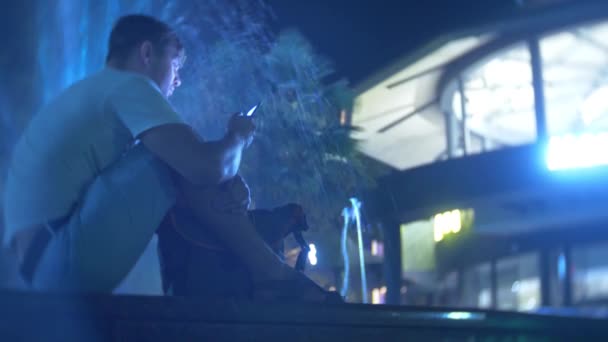 Un joven chico guapo y triste se sienta en la noche al lado de una fuente con iluminación de agua de colores. hablando por teléfono, borroso, 4k — Vídeos de Stock