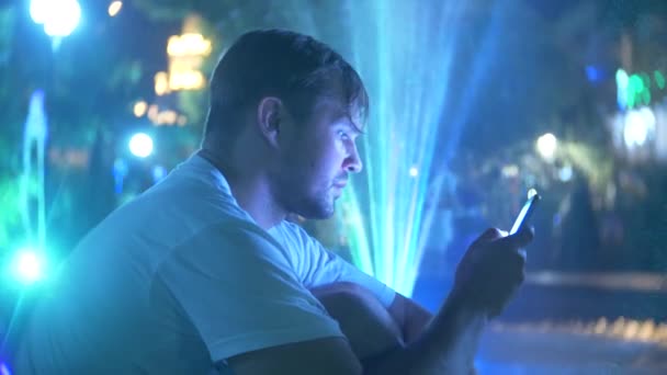 Mladý hezký smutný kluk sedí večer u fontána s osvětlením barevné vody. mluvil po telefonu, rozostření, 4k — Stock video