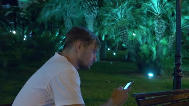 Man zit in de avond in de tropische tuin, onder een palmboom. 4 k. donker — Stockvideo