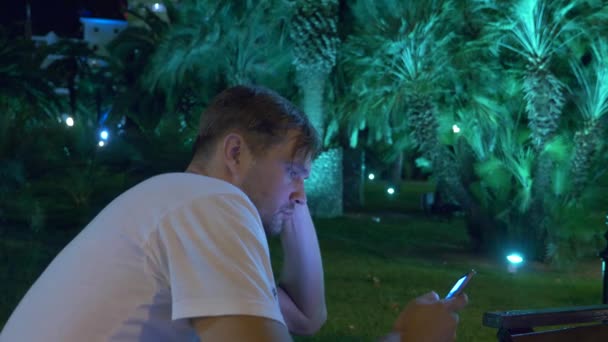 Чоловік сидить увечері в тропічному саду, під пальмою. 4k. темний — стокове відео