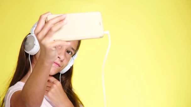 Chica adolescente con auriculares y teléfono móvil escuchando música en el fondo de color. Primer plano, 4k, cámara lenta. hace selfi en el teléfono inteligente — Vídeo de stock