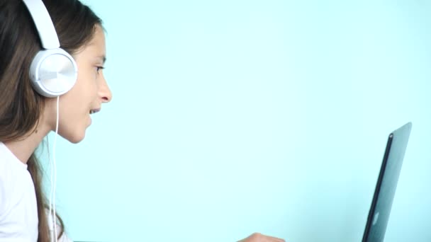 Close-up achtergrond. Een prachtige tienermeisje op een gekleurde achtergrond, in witte koptelefoon kijkt naar het scherm van de laptop. Slowmotion, 4k — Stockvideo