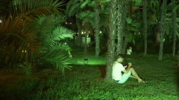 Man zit in de avond in de tropische tuin, onder een palmboom. 4 k. donker — Stockvideo