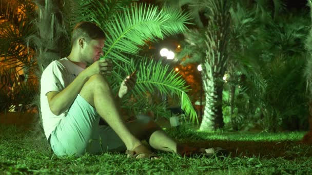 Man sitter på kvällen i den tropiska trädgården, under en Palm. 4 k. mörk — Stockvideo