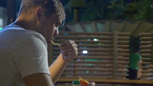 En ledsen person som sitter i en snabbmatsrestaurang utanför på natten äter pommes frites ensam. 4k — Stockvideo