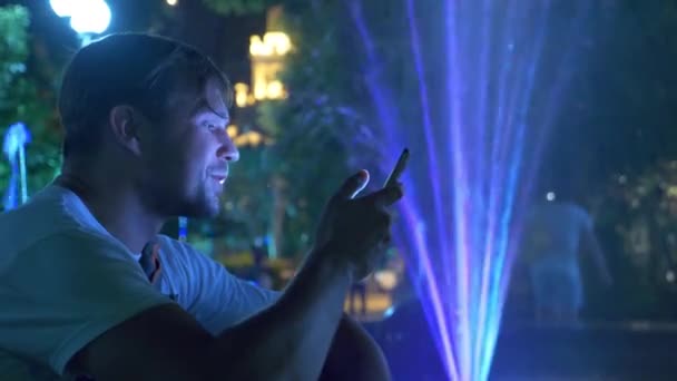 一个年轻英俊的家伙坐在晚上旁边的喷泉与彩色水照明 在电话里聊天 — 图库视频影像
