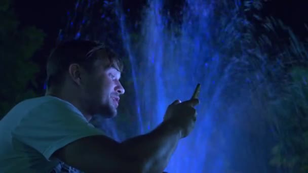 Un chico guapo joven se sienta en la noche al lado de una fuente con iluminación de agua de colores. hablando por teléfono, borroso, 4k — Vídeos de Stock